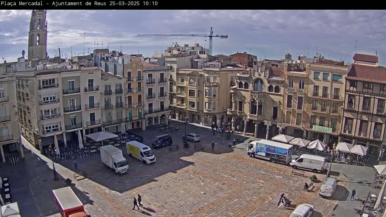 A mostly sunny day reveals a large, cobblestone town square surrounded by multi-story buildings, with several delivery trucks and a few pedestrians present.