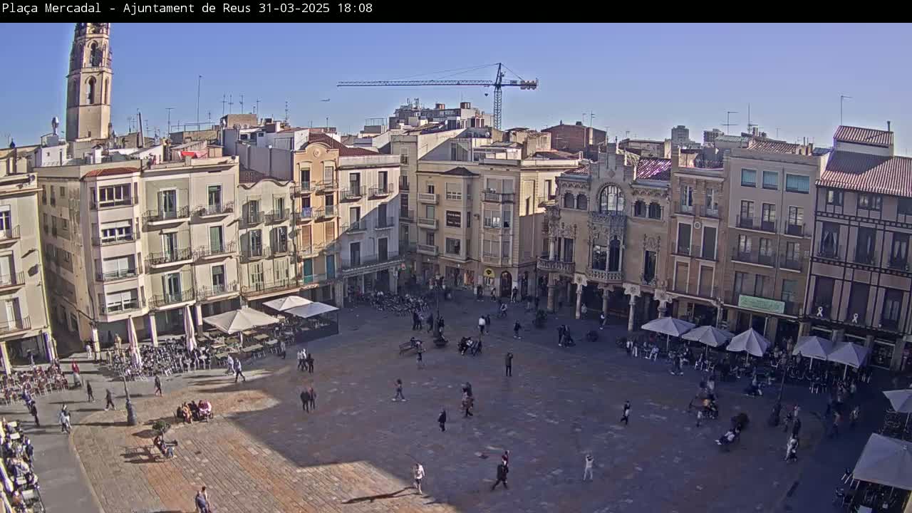 A large town square filled with people and outdoor seating under sunny skies, surrounded by multi-story buildings, including a tall church tower and a construction crane in the distance.