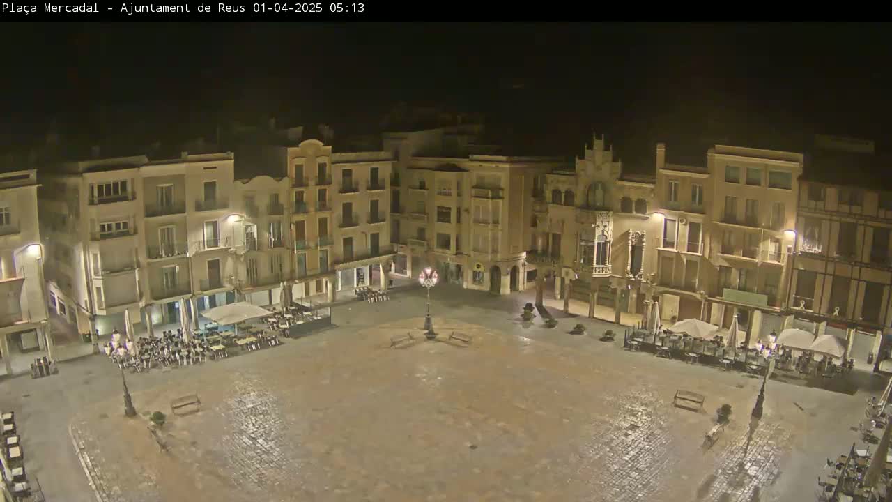 A nighttime, wet, cobblestone town square is surrounded by multi-story buildings with outdoor seating areas.