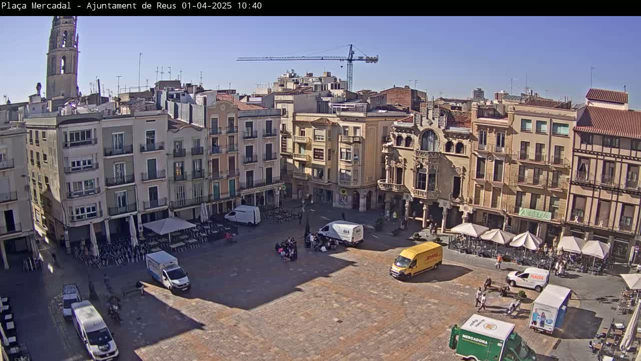 A sunny city square with several buildings surrounding it, a few parked vans, and some people gathered in the center.