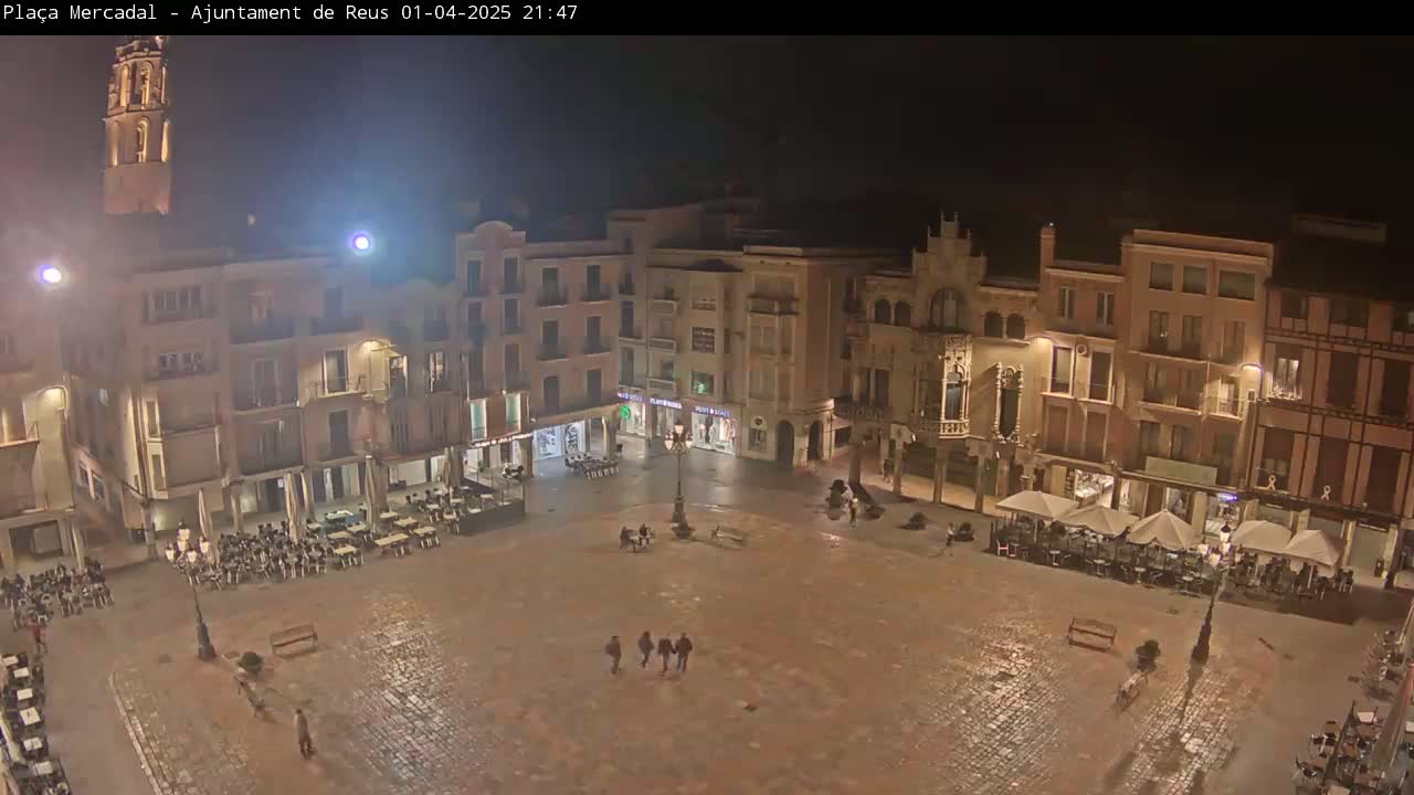 A nighttime, high-angle view of a mostly empty town square, with several buildings surrounding it and a few people walking.