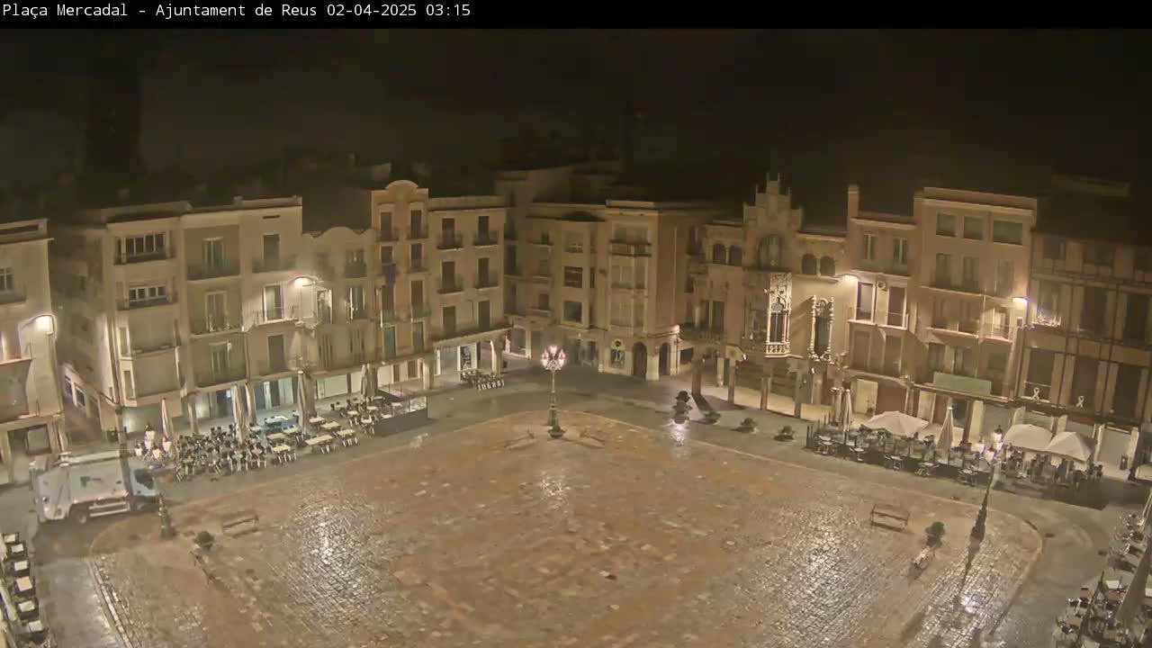 A rain-slicked, empty town square at night, surrounded by multi-story buildings with illuminated windows and sparsely occupied outdoor seating areas.