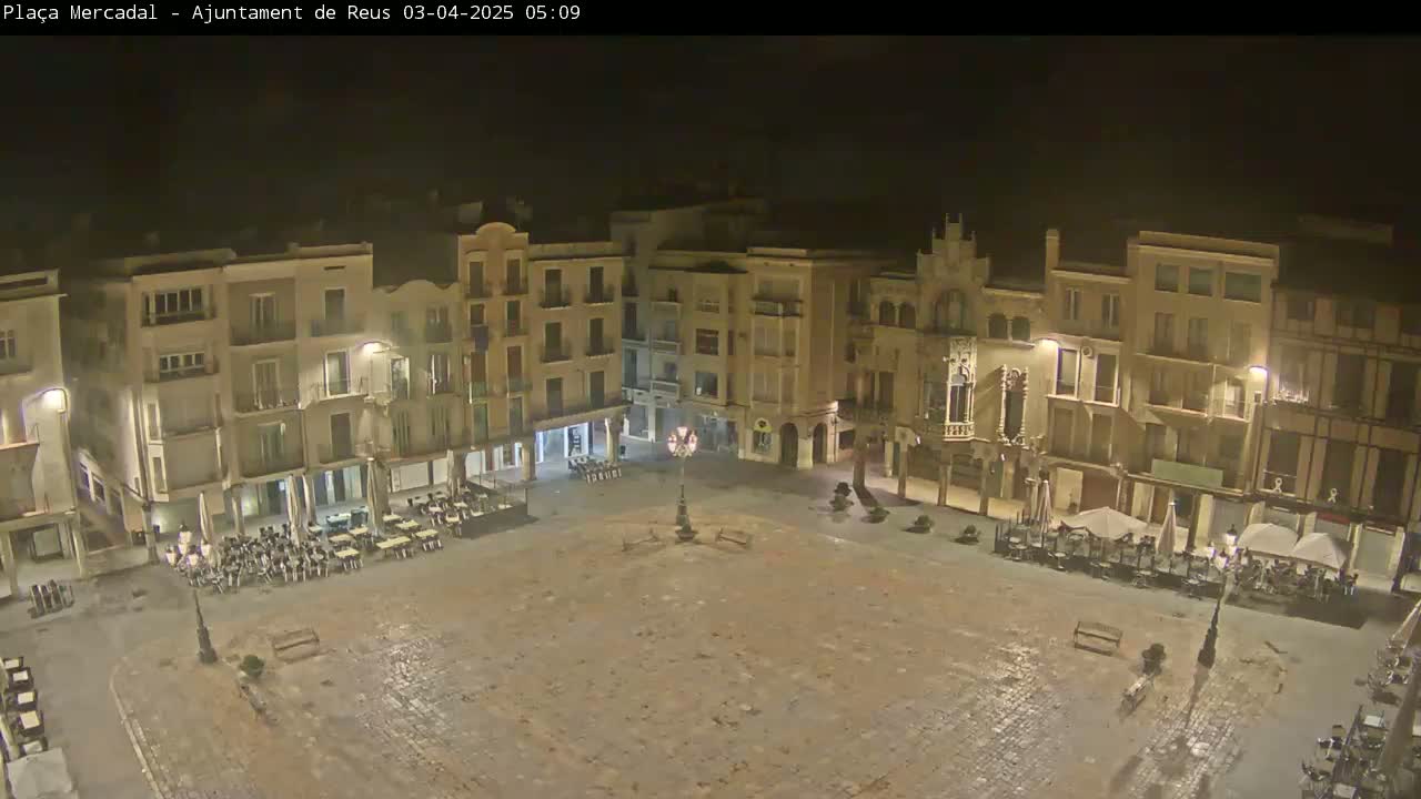 A mostly empty town square at night, surrounded by multi-story buildings with outdoor seating areas, is lightly illuminated.