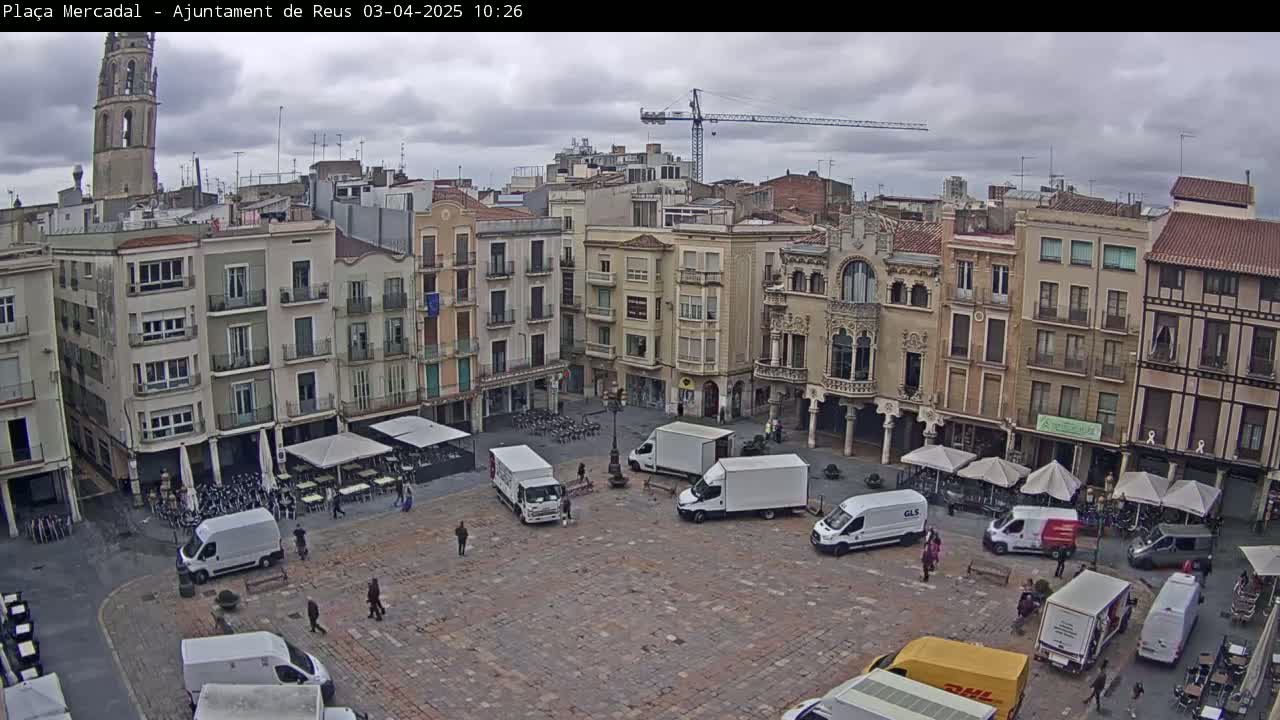 An overcast day reveals a town square filled with delivery vans, outdoor seating, and several multi-story buildings surrounding it, one of which has a tall tower.