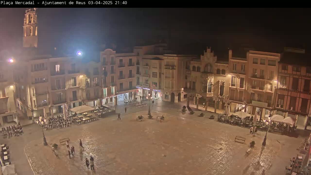 A nighttime view of a town square, surrounded by buildings with lights illuminating the wet cobblestone surface.