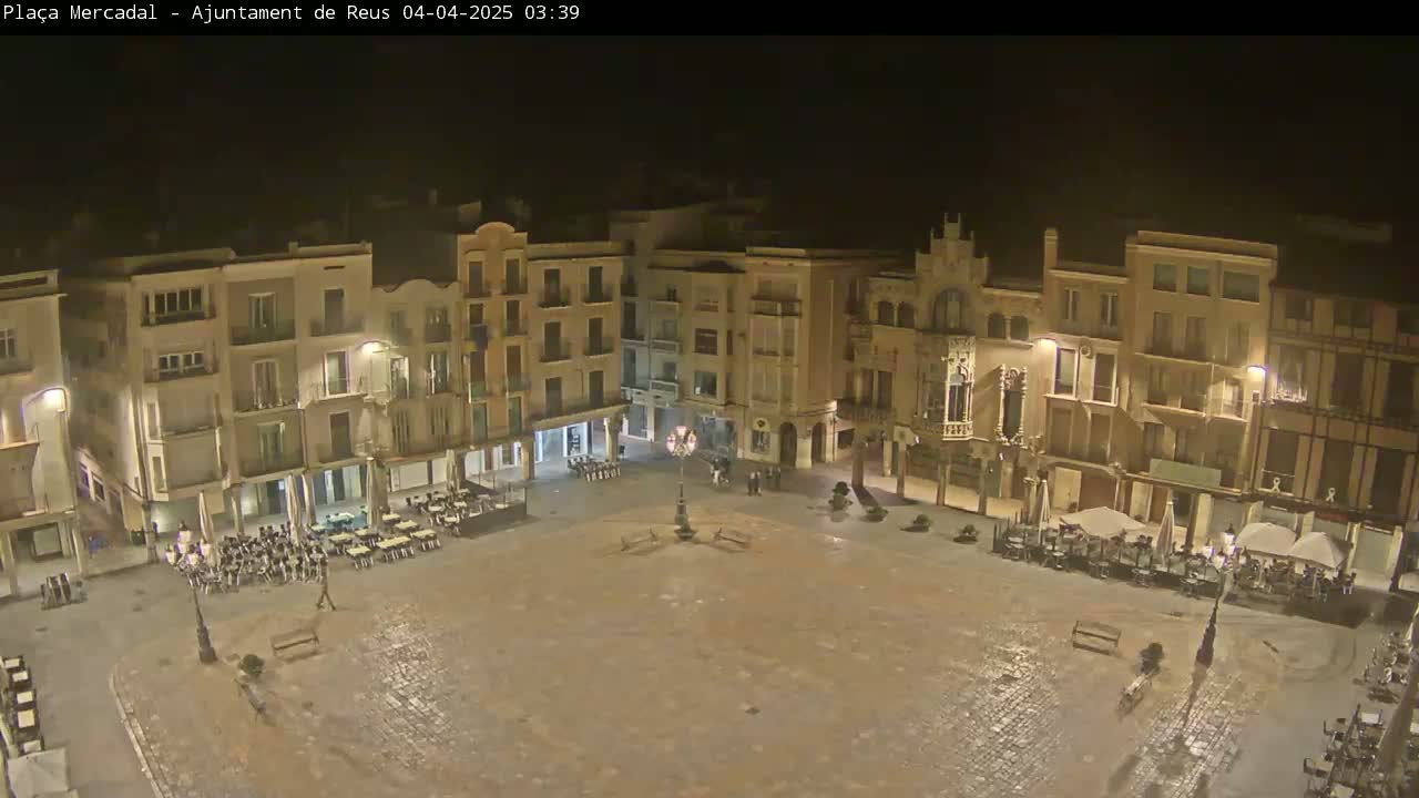 A mostly empty town square at night, with several buildings surrounding it and some outdoor seating areas, is slightly wet.