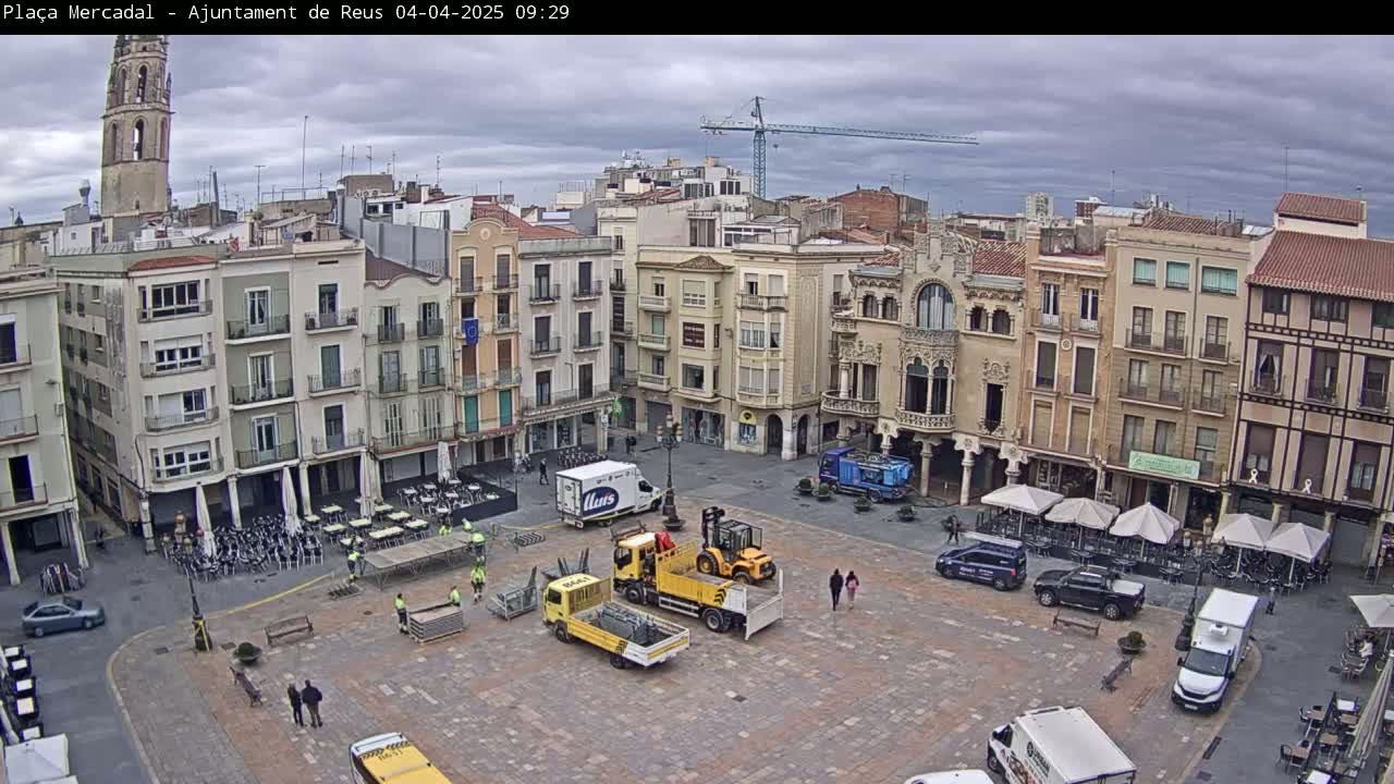 A large town square, under an overcast sky, is being prepped for an event with several trucks and pieces of equipment scattered across the stone pavement;  buildings surround the square, and a tall tower is visible in the distance.
