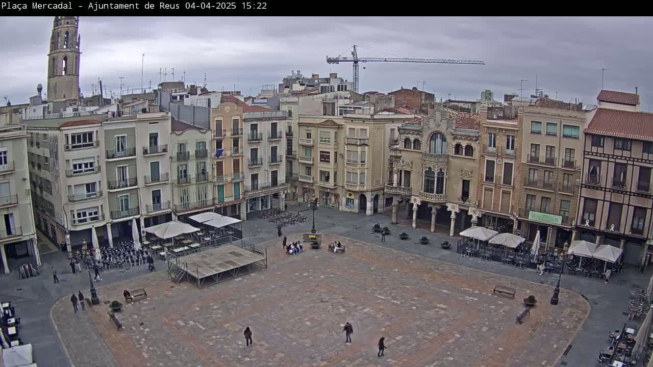 A large, mostly empty town square with surrounding buildings under an overcast sky, featuring a construction crane in the background.