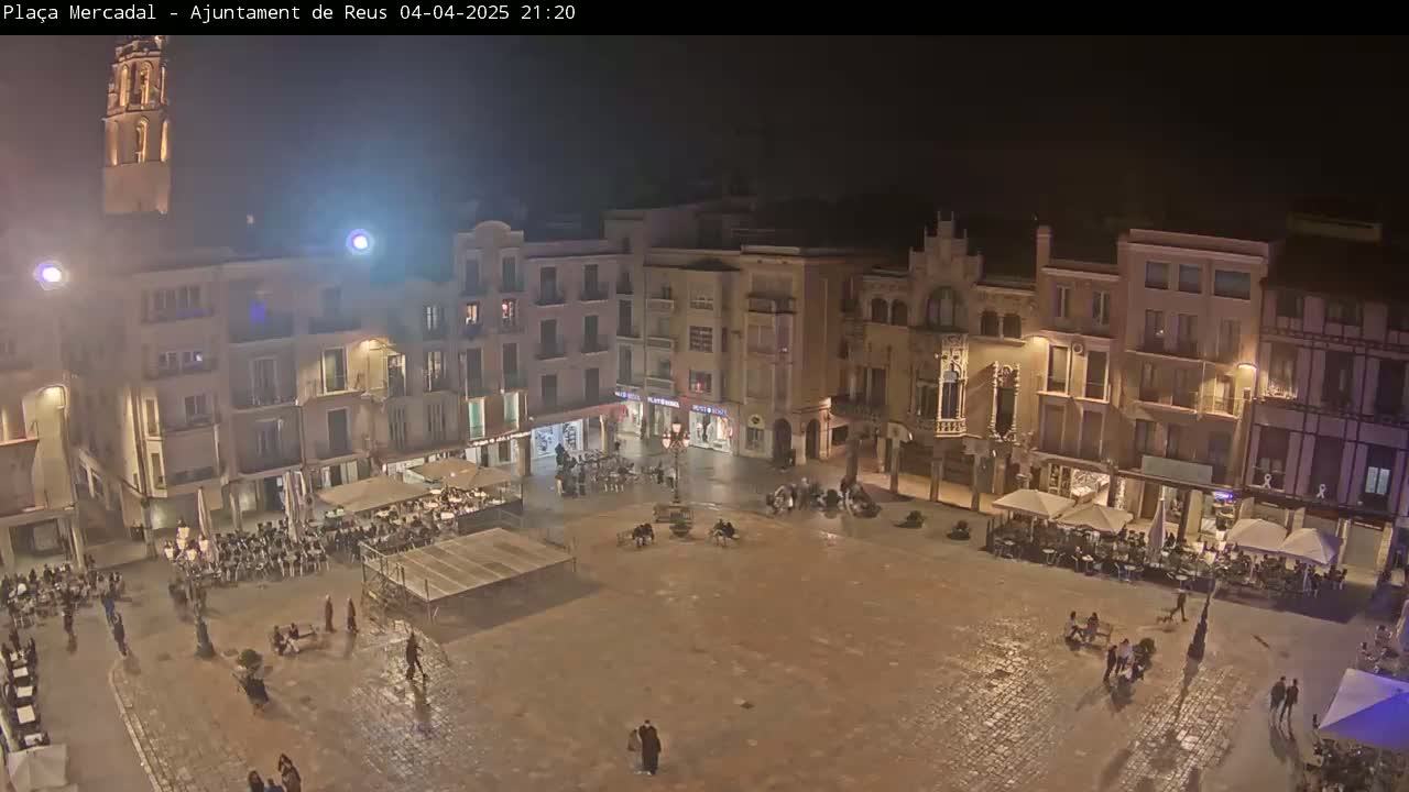 A nighttime, wet town square is surrounded by buildings with lights on, and people are sitting at outdoor cafes and walking around.