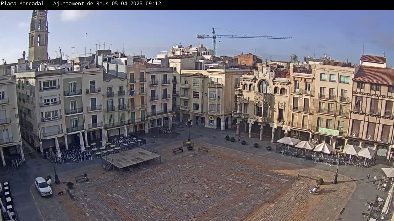 A large, cobblestone town square is surrounded by multi-story buildings under a clear, sunny sky, with a few people and outdoor seating at cafes visible.