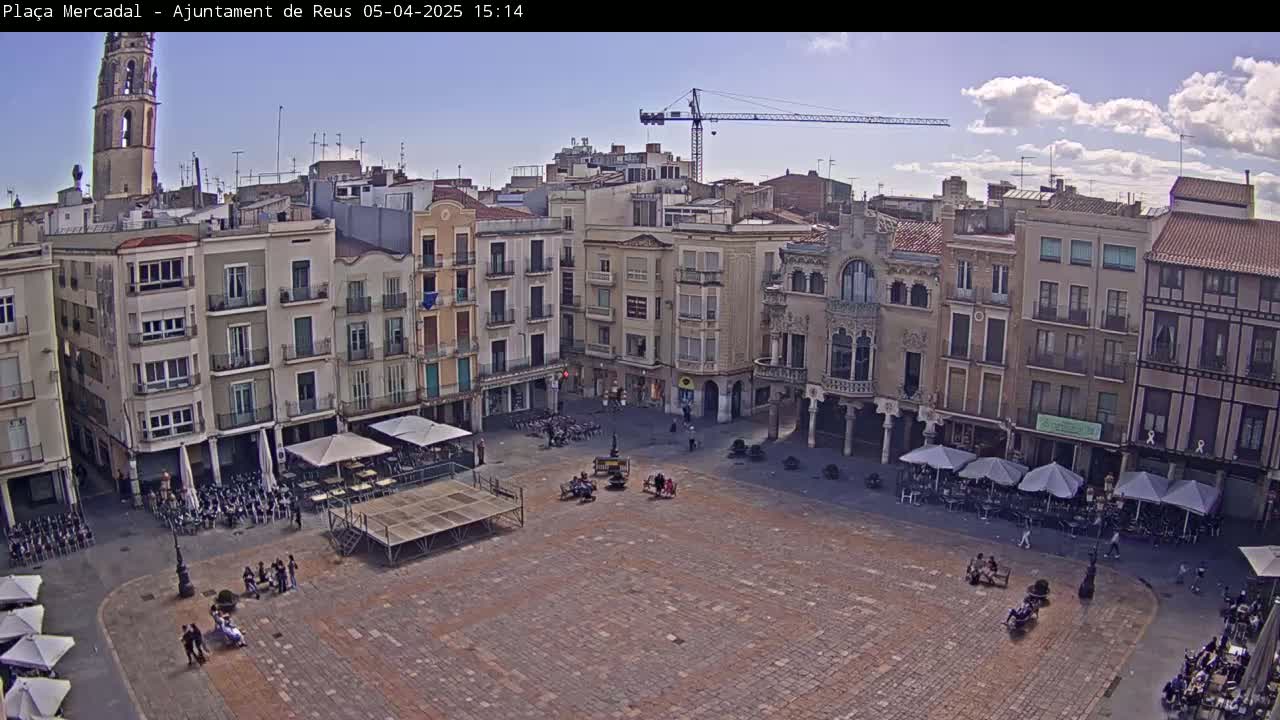 A partly cloudy day reveals a large, paved town square surrounded by multi-story buildings, with several outdoor cafes and a small number of people present.