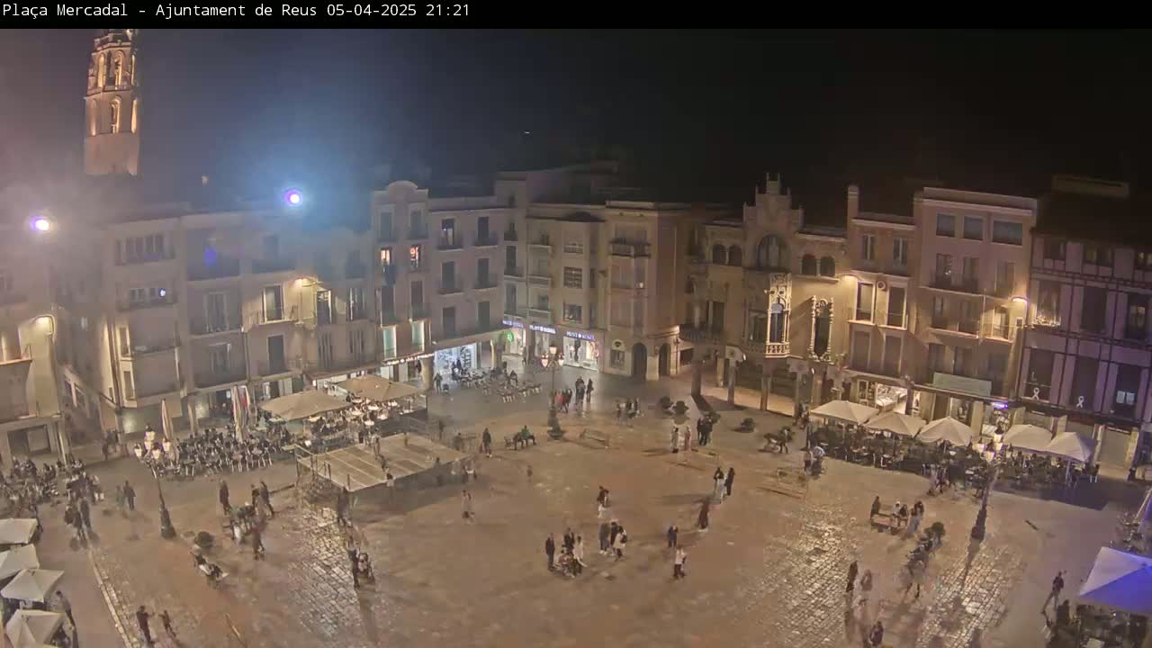 A nighttime view of a town square, under a dark sky, shows several people walking around the cobblestone plaza with outdoor seating at restaurants and cafes surrounding the square's perimeter.