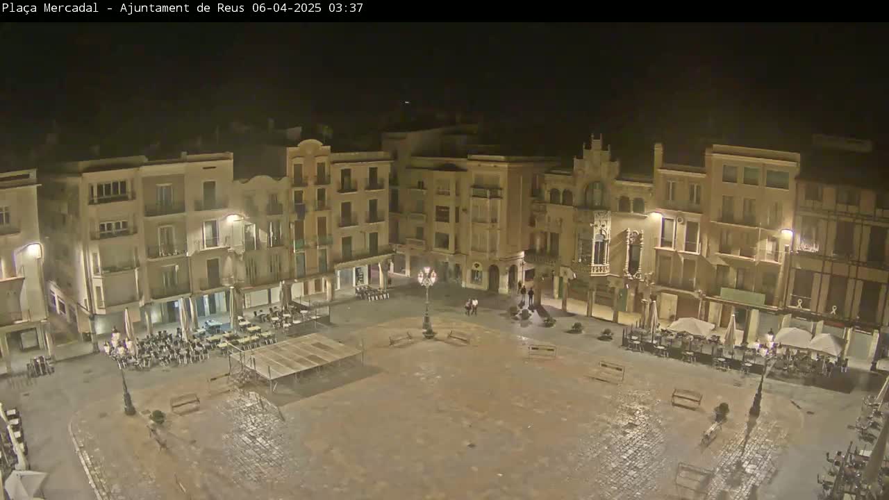 A nighttime, overhead view of a town square, mostly empty except for a few people and some outdoor seating at cafes lining the square's perimeter.
