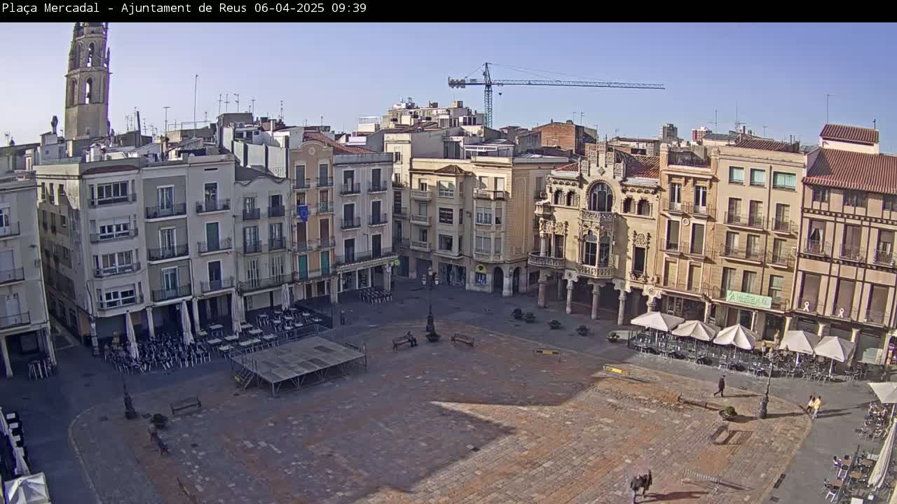 A large, mostly empty town square with surrounding buildings, a few people, and outdoor seating under umbrellas, all under a clear sunny sky.
