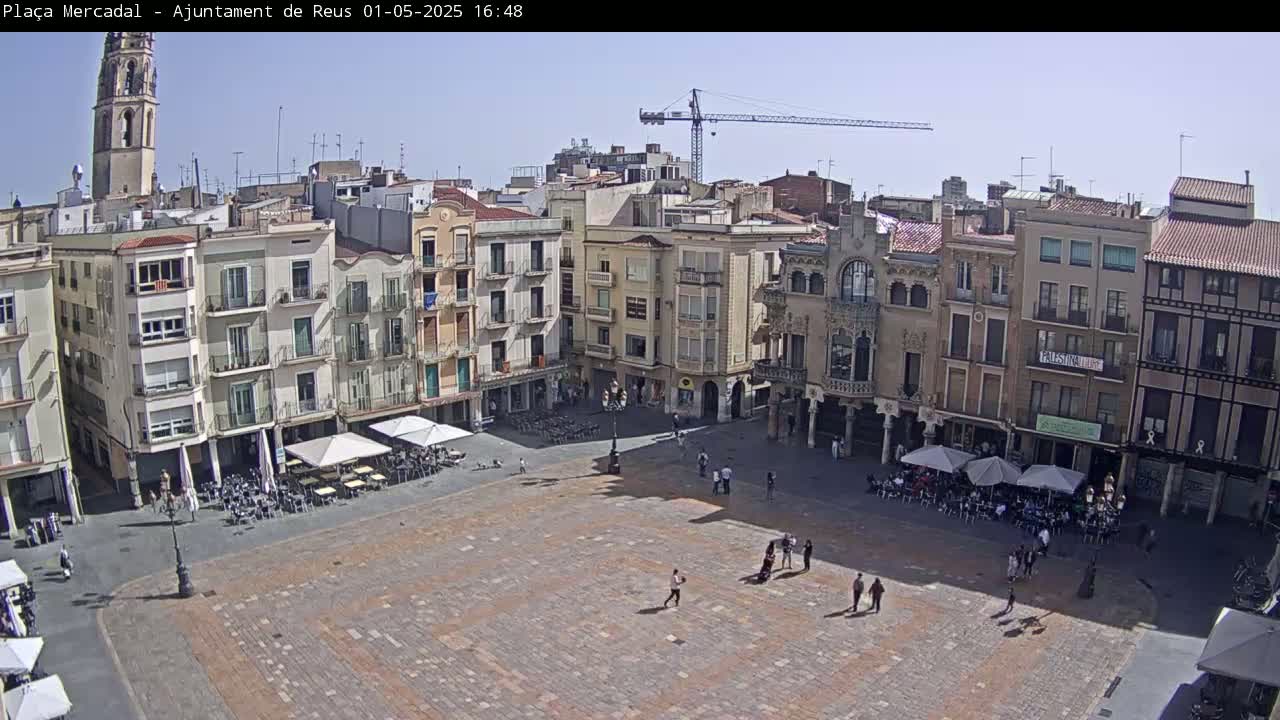 A large, paved town square is filled with sparsely scattered people and outdoor cafe seating under sunny skies, surrounded by multi-story buildings, one of which has a tall tower.
