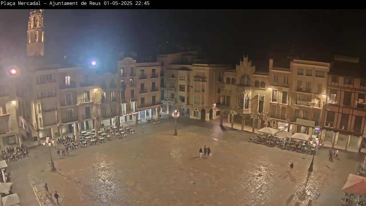 A nighttime view of a town square, illuminated by streetlights, with several people and outdoor seating areas around the perimeter of the square, wet from recent rain.
