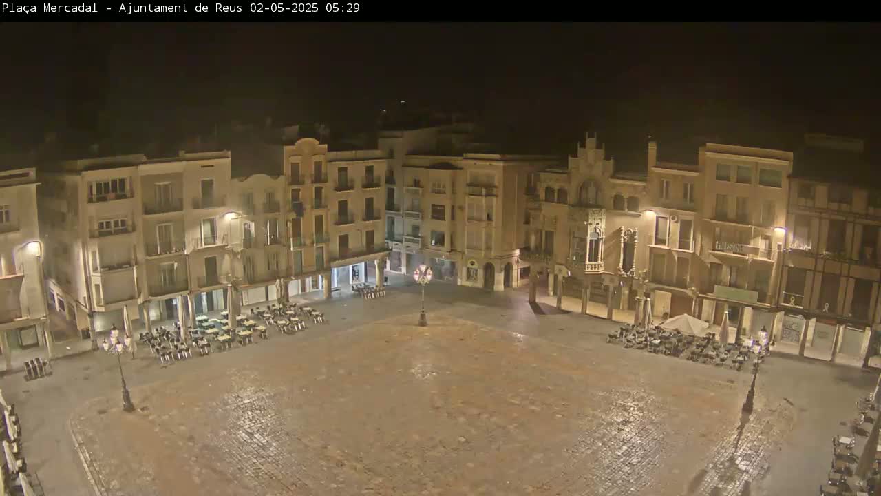 A nighttime, wet, cobblestone town square is surrounded by multi-story buildings with outdoor seating areas.