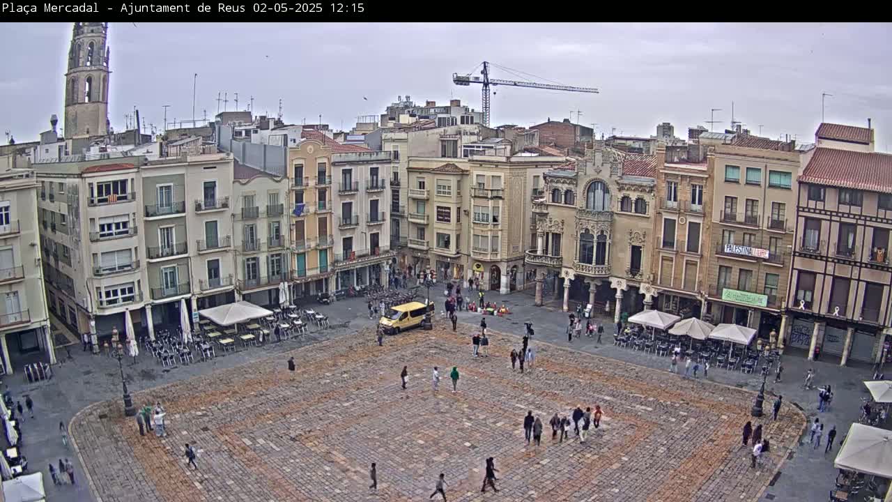 A large, paved town square, surrounded by multi-story buildings, with a few people and outdoor seating areas under cloudy skies.