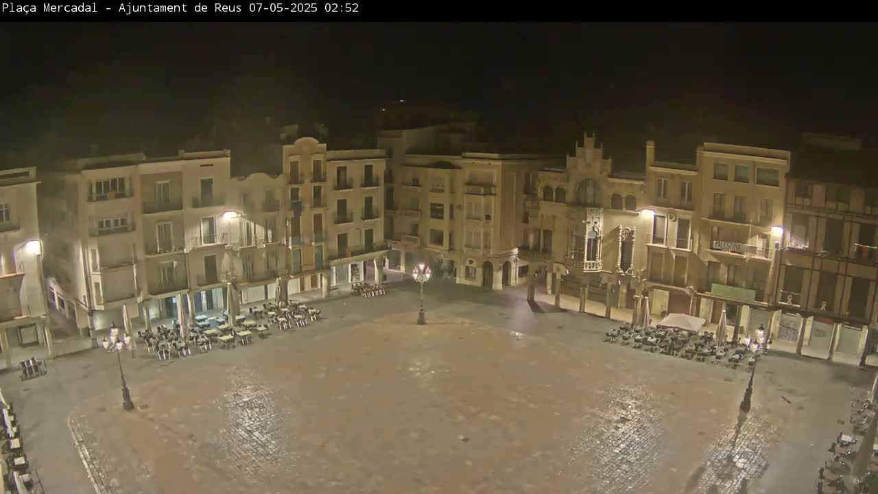 A mostly empty plaza at night, surrounded by multi-story buildings, is lightly wet from a recent rain.