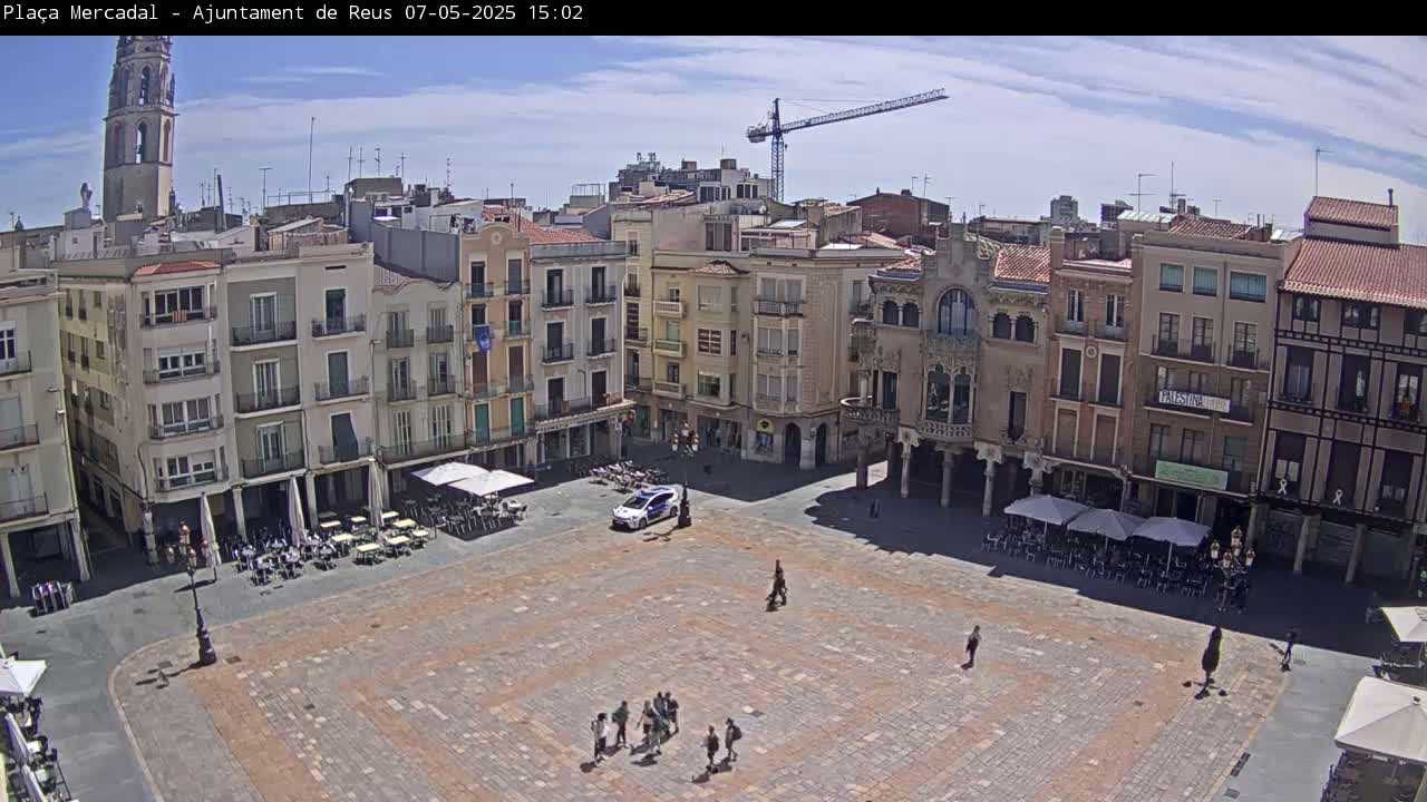 A large, paved town square is surrounded by multi-story buildings under a mostly sunny sky, with a few people and outdoor seating areas visible.