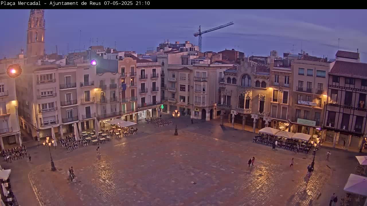 A town square at dusk is sparsely populated with people sitting at outdoor cafes under the dim light of the buildings surrounding it.