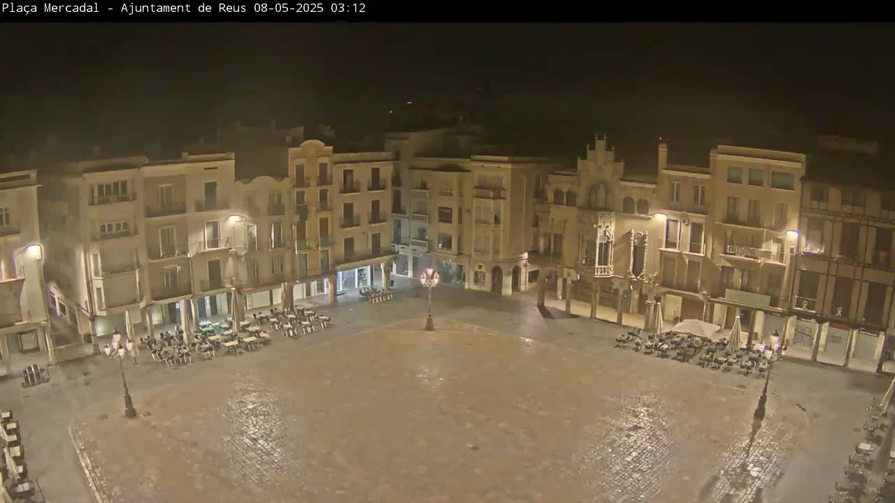 A mostly empty cobblestone plaza at night, surrounded by multi-story buildings, is lightly wet from recent rain.