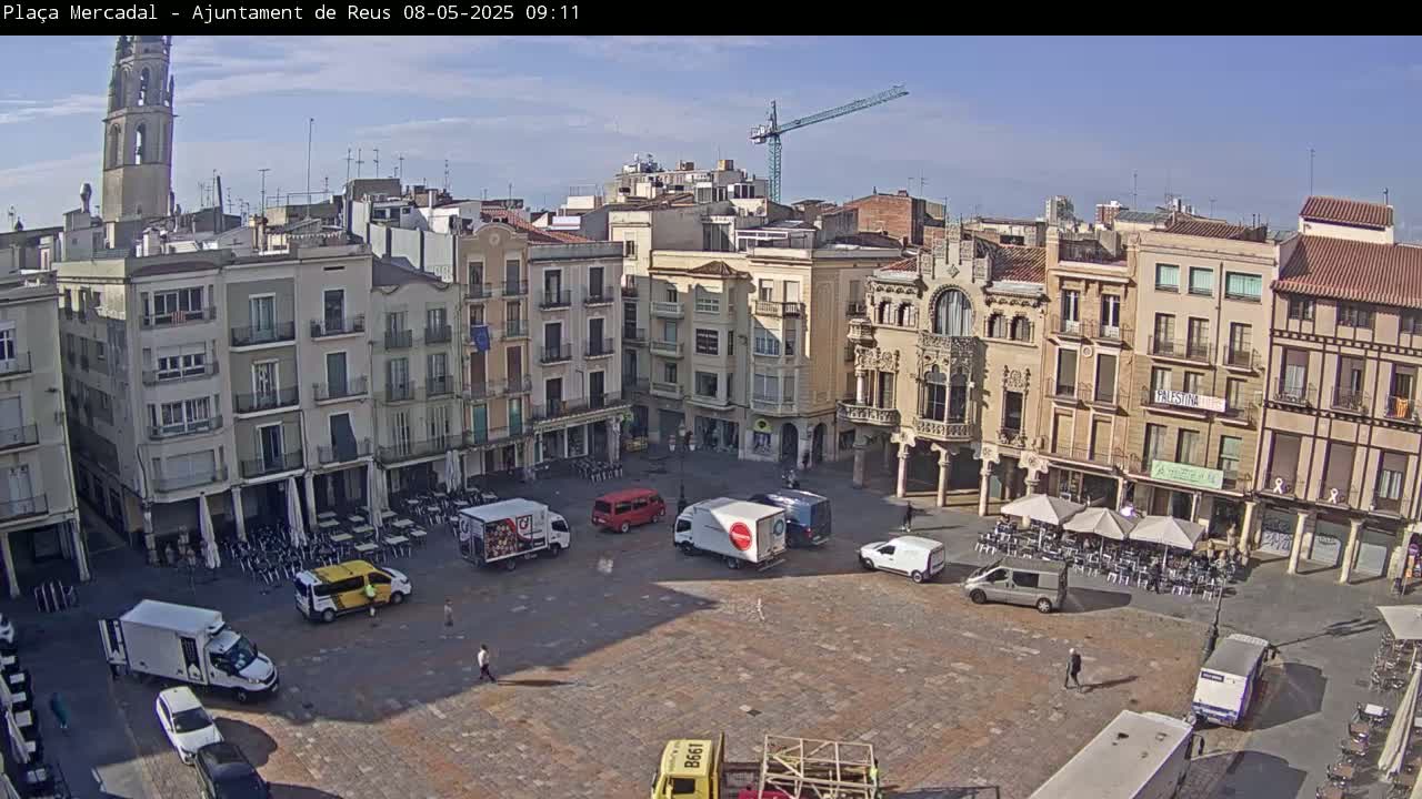 A sunny day reveals a large town square surrounded by multi-story buildings, with several vehicles and outdoor seating areas present.