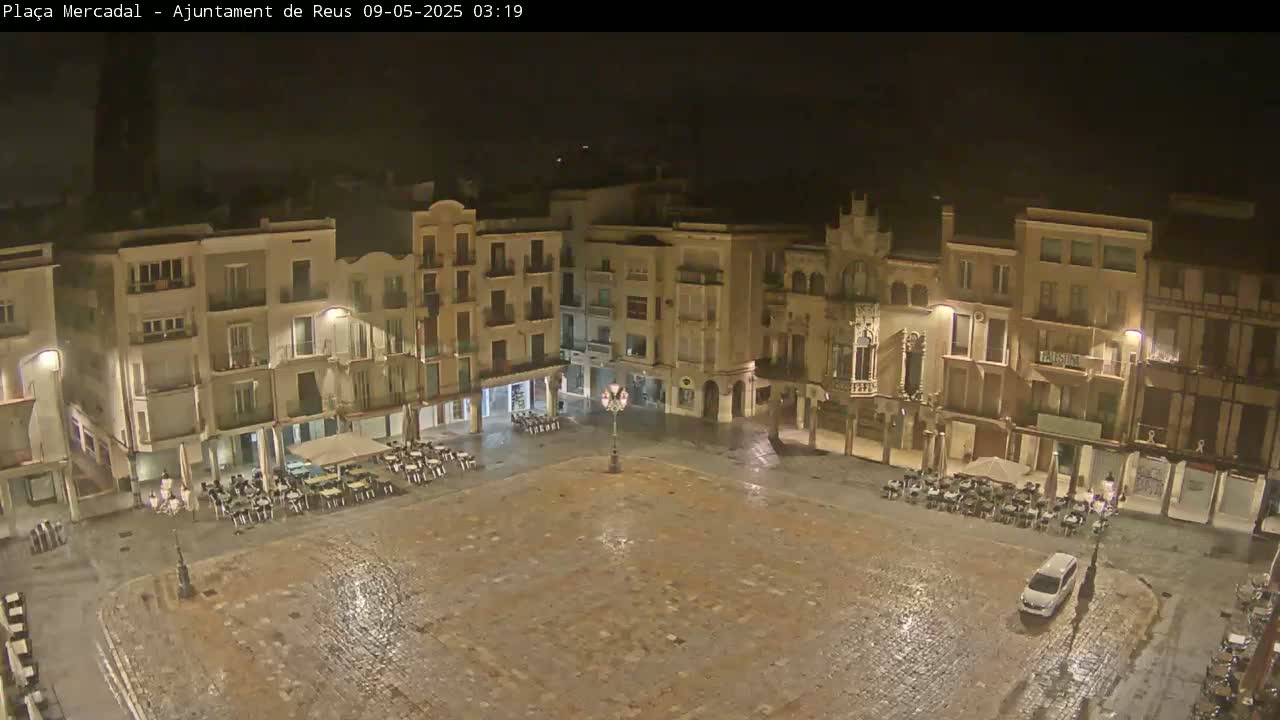 A rain-slicked, empty town square at night is surrounded by multi-story buildings with lights illuminating the scene.