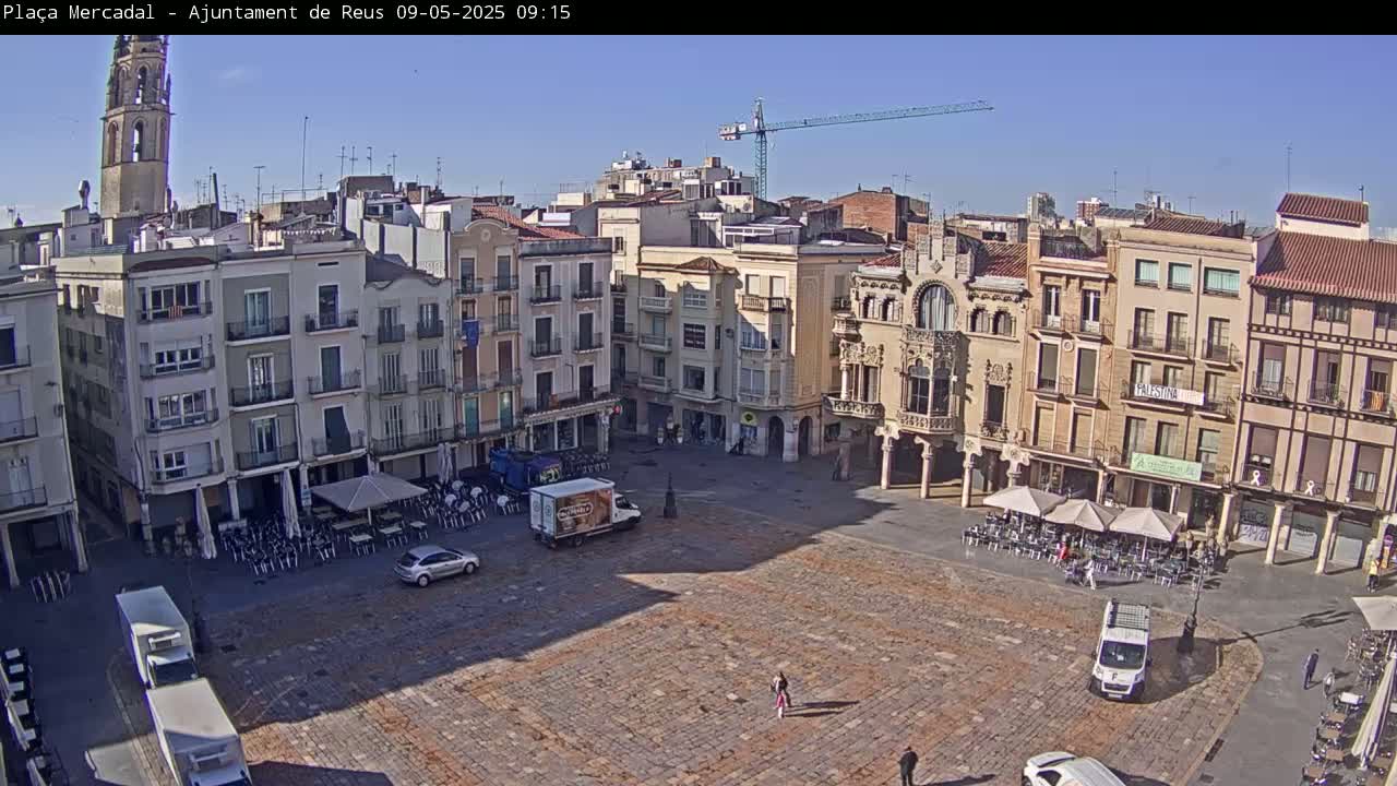 A large, mostly empty plaza in a town, surrounded by multi-story buildings under a clear, sunny sky, with a few pedestrians and parked vehicles visible.