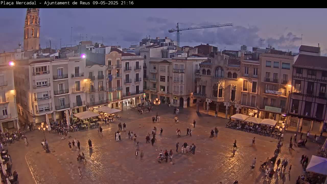 A town square at dusk, under a partly cloudy sky, is populated with people and outdoor seating areas at various establishments surrounding the square.
