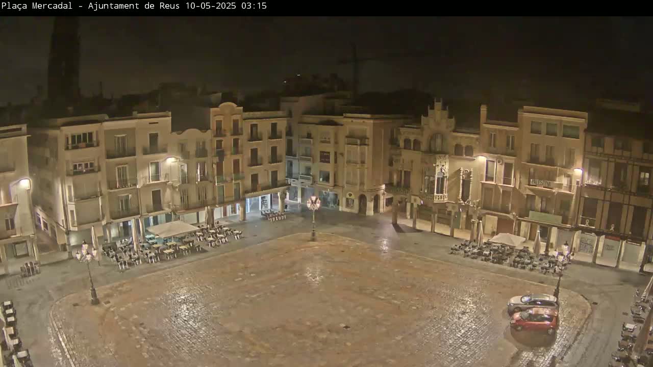 A wet, empty town square at night, surrounded by multi-story buildings with outdoor seating areas.