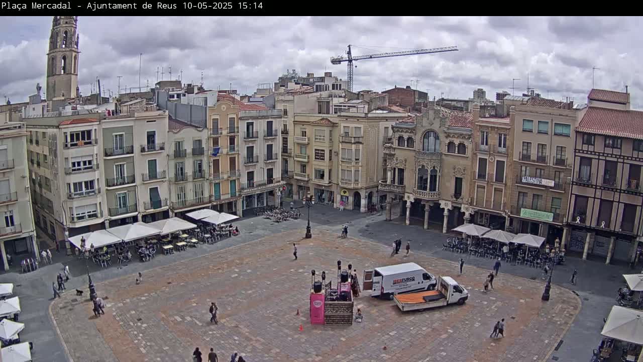 A large town square with several buildings surrounding it, under a partly cloudy sky, features outdoor cafes and a small crowd of people.
