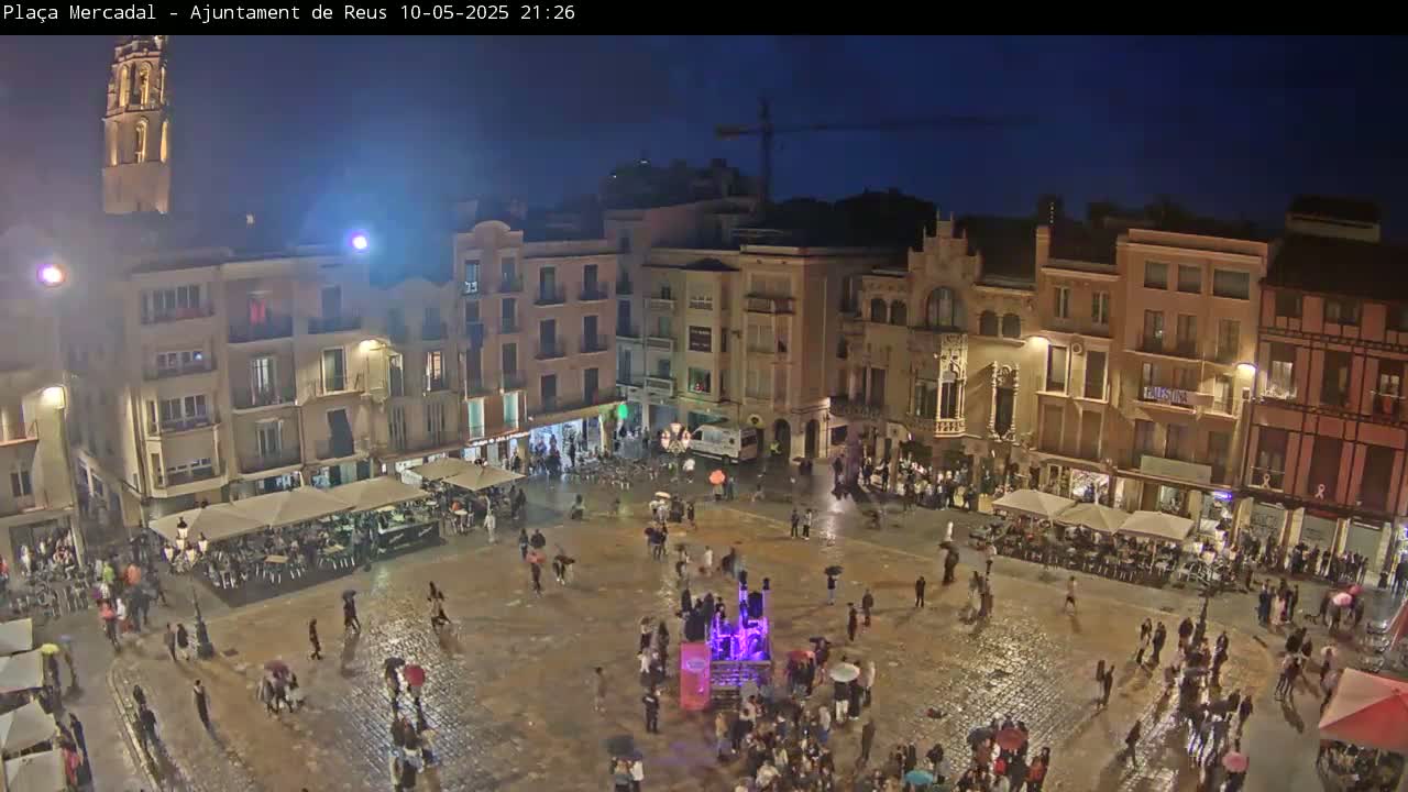 A rain-slicked town square at night is filled with people under the glow of streetlights and building lights, surrounded by multi-story buildings.