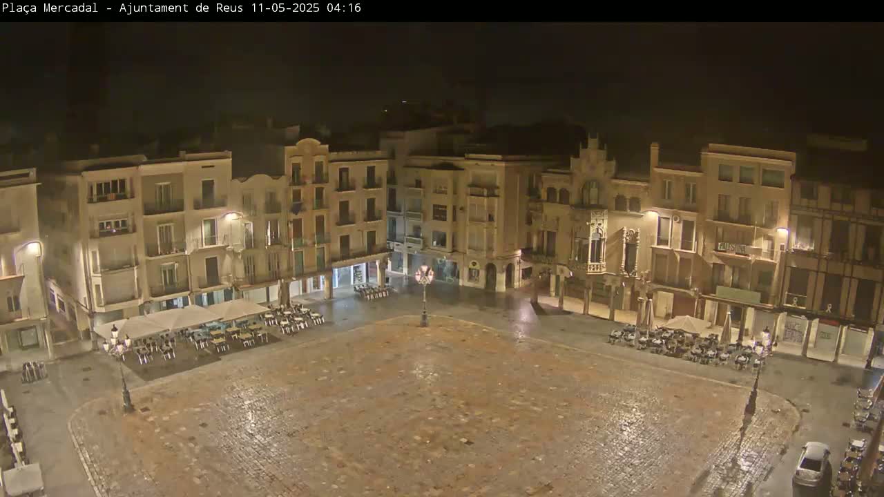 A wet, empty town square at night is surrounded by multi-story buildings with outdoor seating areas under umbrellas.