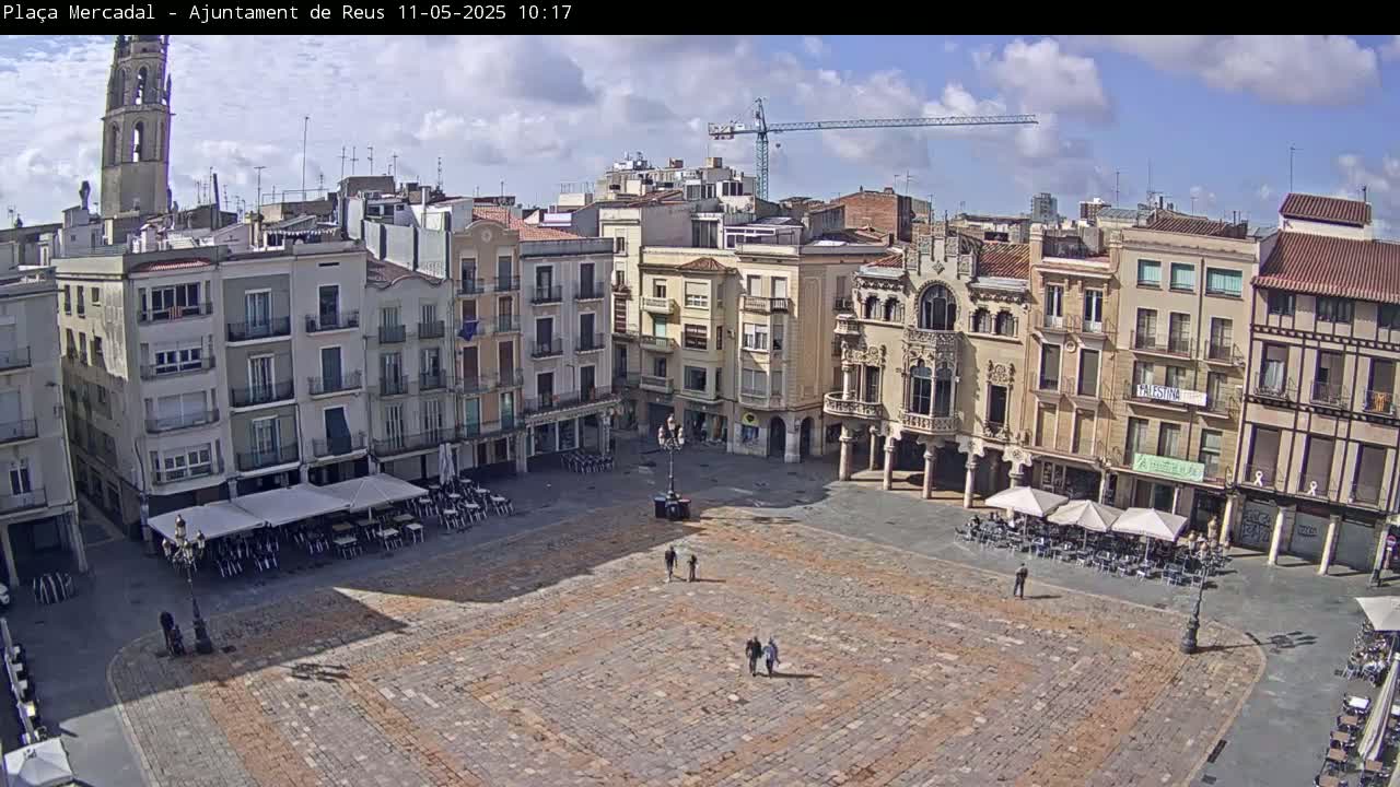 A mostly empty cobblestone town square is surrounded by multi-story buildings under a partly cloudy sky.