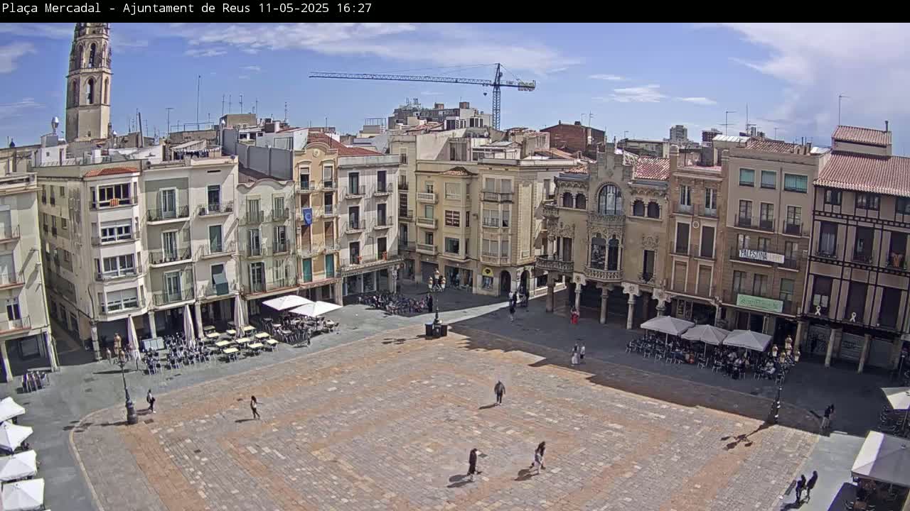 A mostly empty town square with surrounding buildings, outdoor seating areas, and a construction crane visible in the distance under a mostly sunny sky.