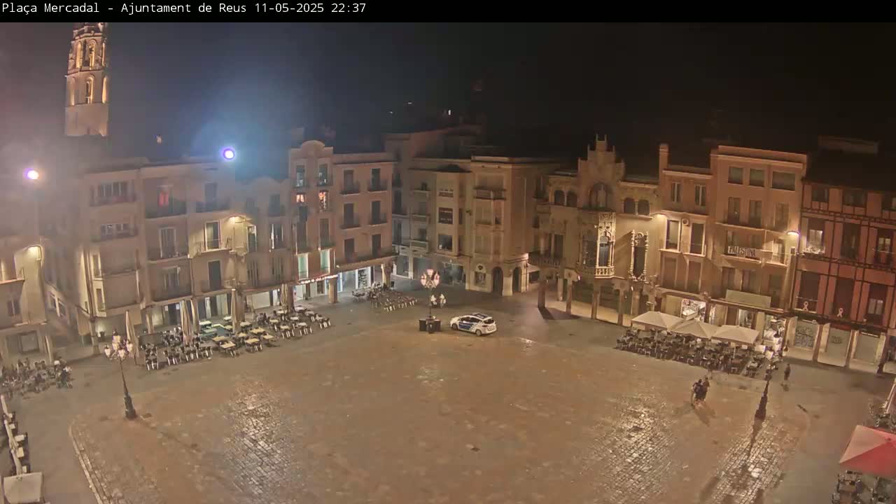 A nighttime view of a town square with several buildings surrounding it, outdoor seating areas, and a single vehicle parked in the center.