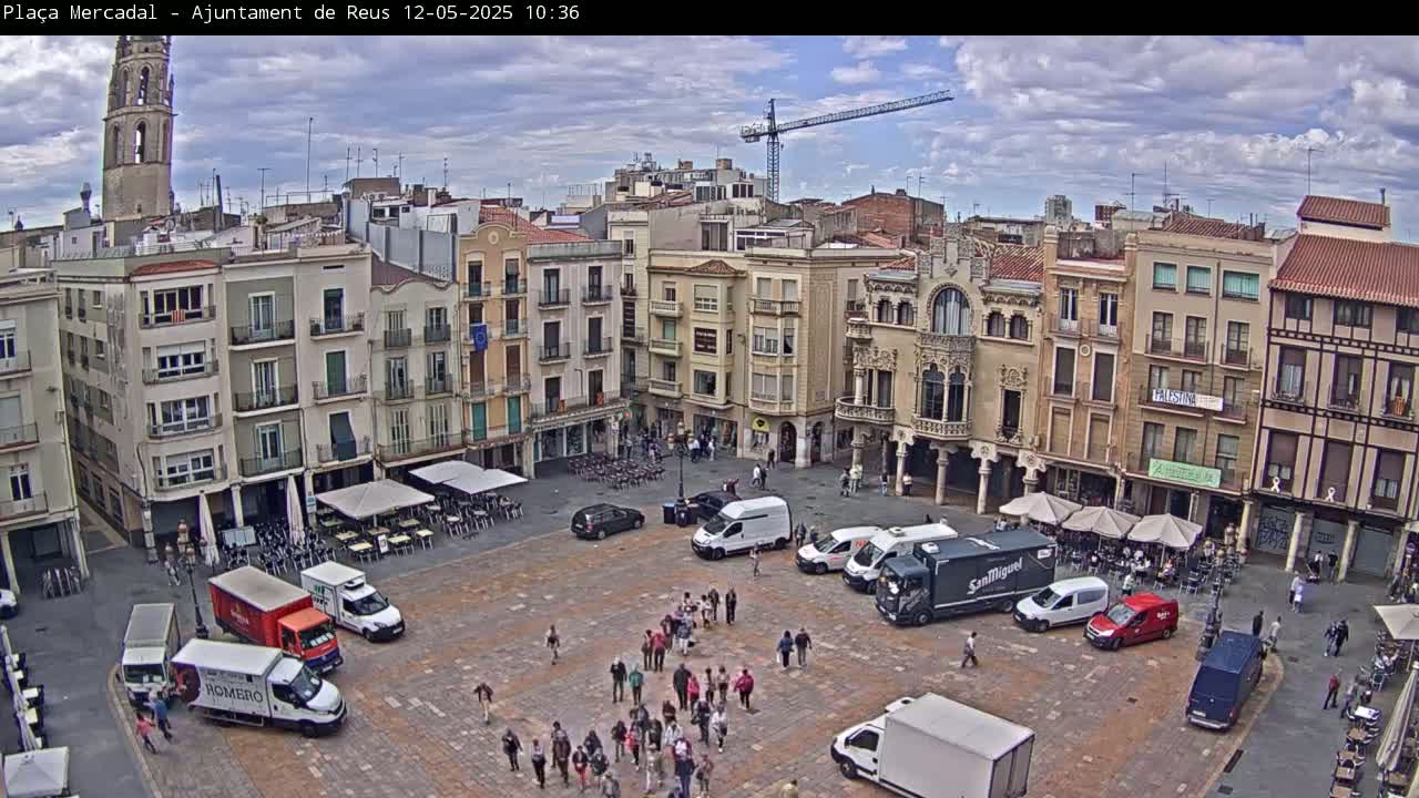 A mostly sunny, partly cloudy day reveals a large town square filled with people, vehicles, and outdoor seating areas, surrounded by multi-story buildings, one of which is a tall church tower.