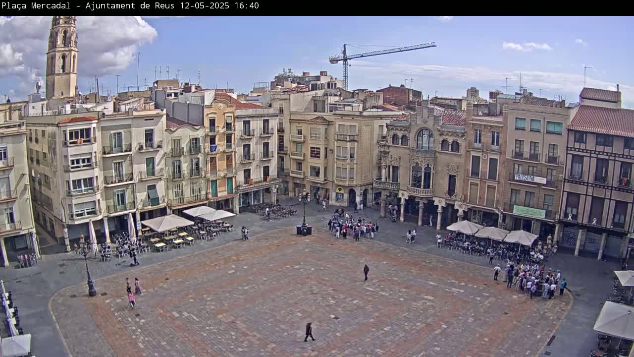 A mostly sunny day shows a large, paved town square surrounded by multi-story buildings, with people gathered in small groups and seated at outdoor cafes.