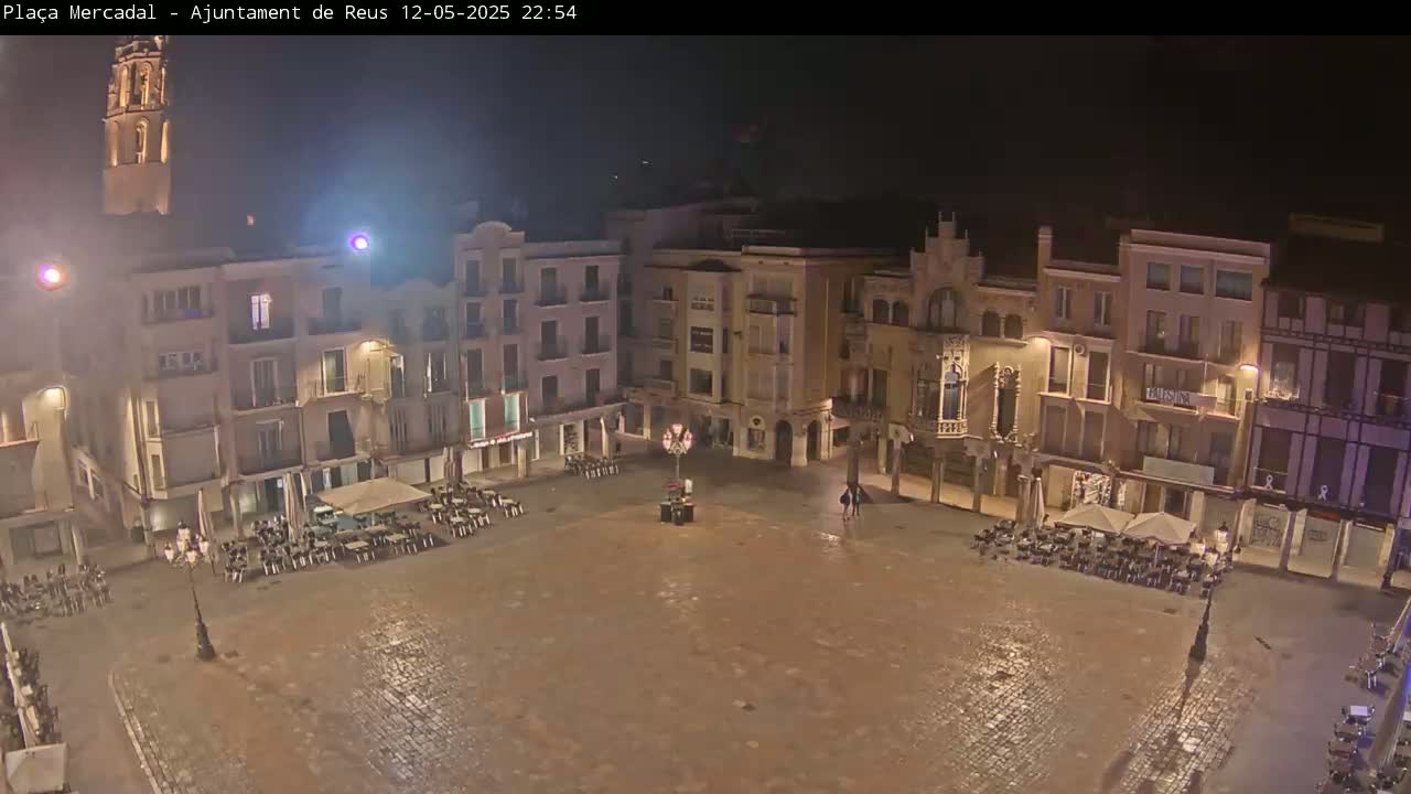 A mostly empty, cobblestone town square at night is surrounded by multi-story buildings with some outdoor seating areas.