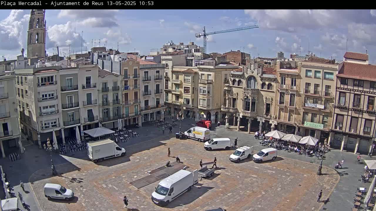 A mostly sunny town square, filled with several vans and people sitting at outdoor cafes, is surrounded by multi-story buildings, one of which has a tall tower.