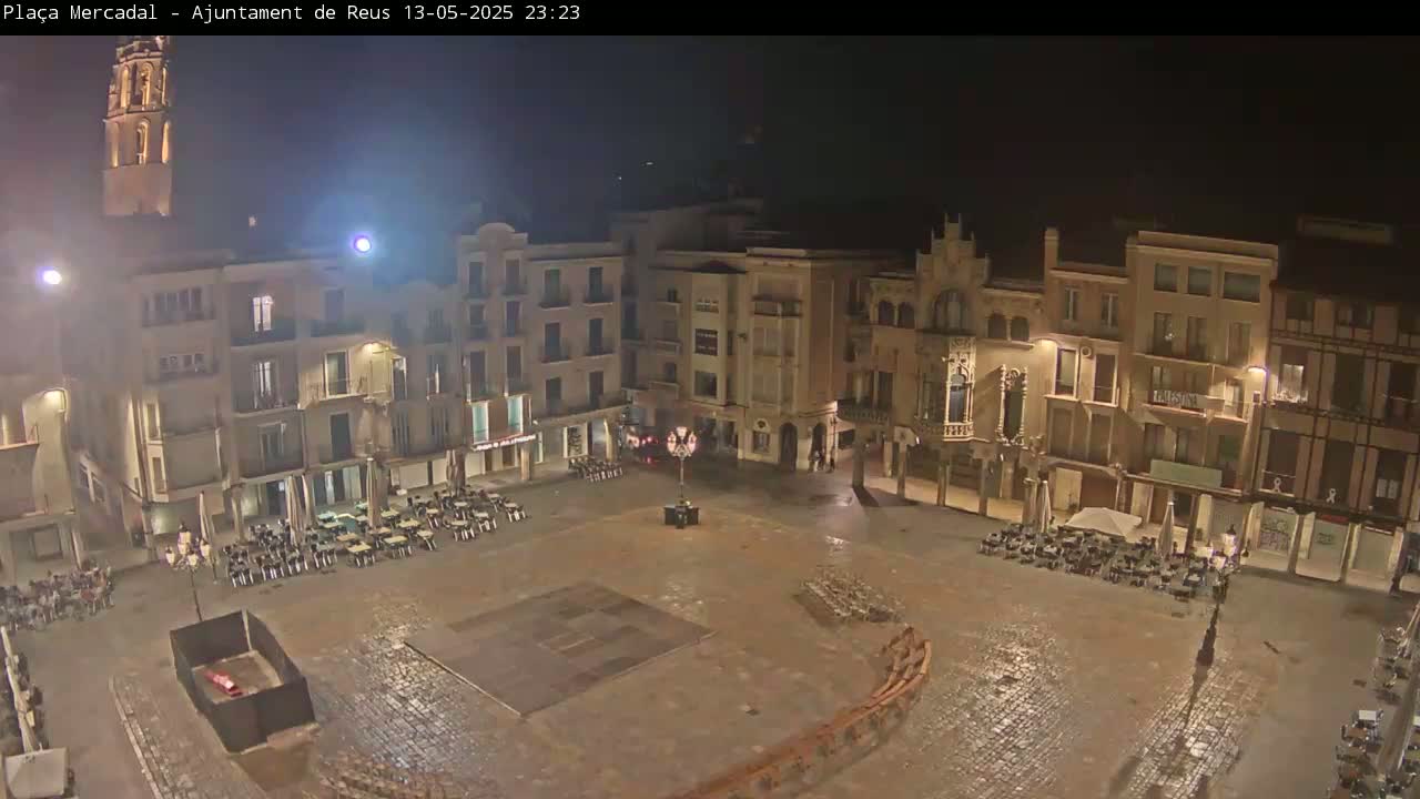 A nighttime view of a town square, surrounded by buildings, with outdoor seating areas and a mostly empty, paved center.