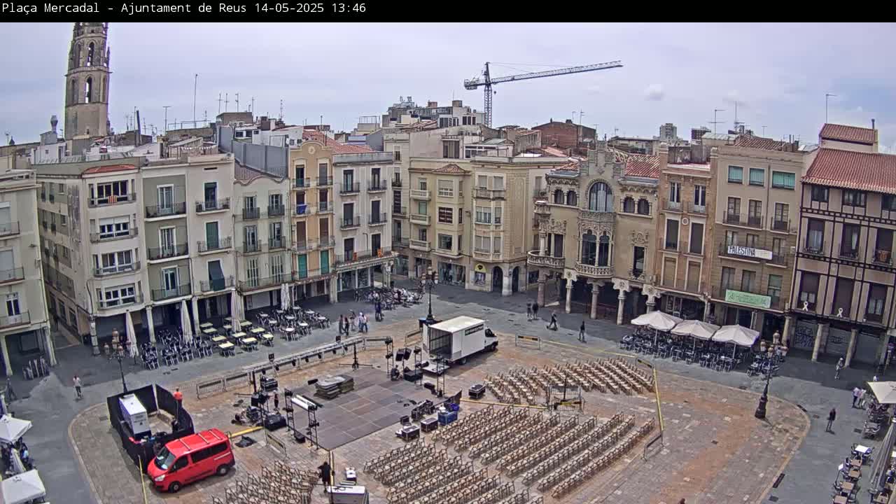 A mostly sunny town square is set up for an outdoor event, with rows of chairs and a stage in the center, surrounded by multi-story buildings.