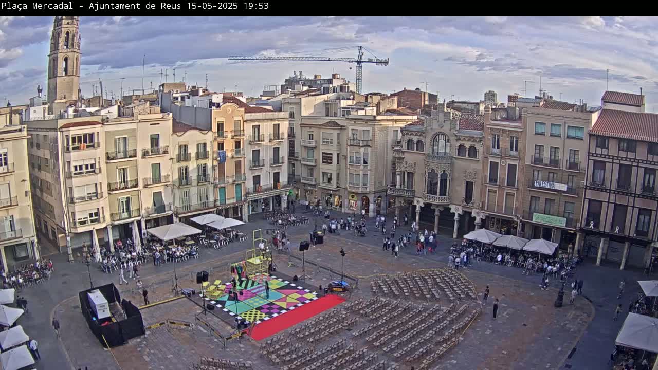 A town square with numerous buildings surrounding it, a stage with colorful geometric designs in the center, and many chairs set up, under a partly cloudy sky.
