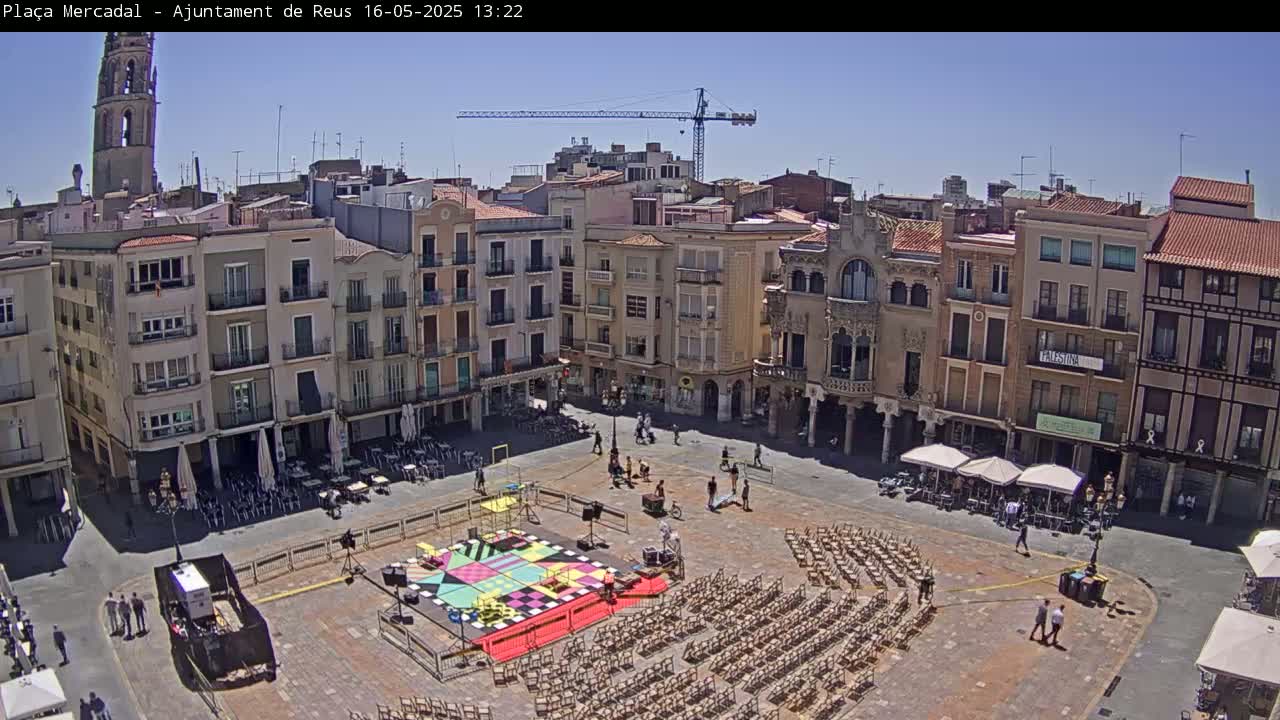 A town square with multiple-story buildings surrounding it is under a sunny sky; the square contains many chairs set up, a colorful design painted on the ground, and several people scattered about.
