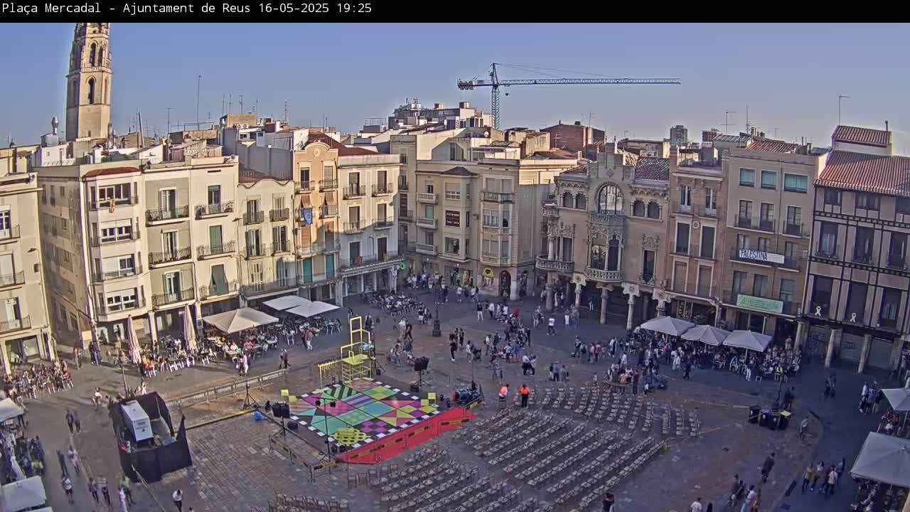 A large town square filled with people, featuring a colorful stage setup, numerous chairs, and surrounding buildings under a clear, sunny sky.