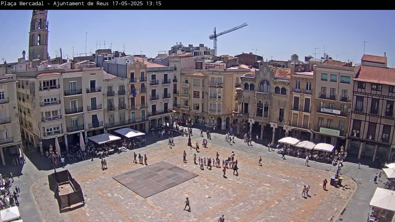 A sunny day reveals a large town square surrounded by multi-story buildings, with people scattered throughout the plaza and outdoor seating at cafes.