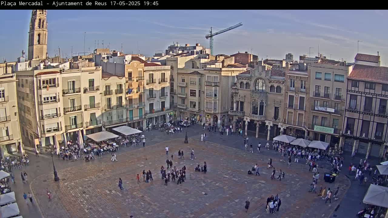 A large town square, populated with people and outdoor seating, is surrounded by multi-story buildings under a clear, pale blue sky.