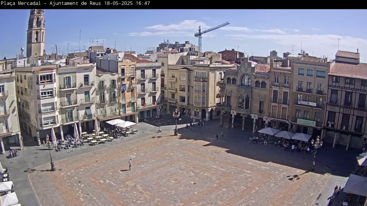 A mostly empty town square with surrounding buildings under a partly sunny sky, features outdoor seating at various cafes and a construction crane in the background.
