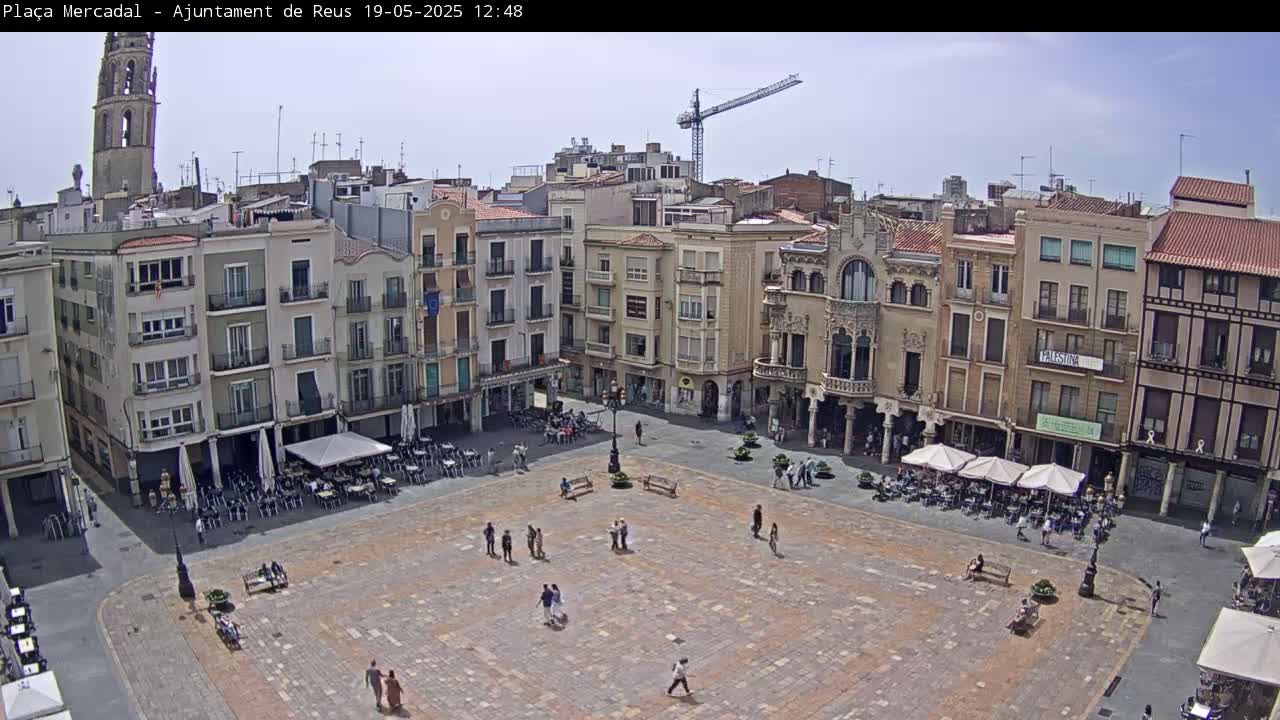 A large, mostly empty town square with several people and outdoor cafes is surrounded by multi-story buildings under a clear sky.